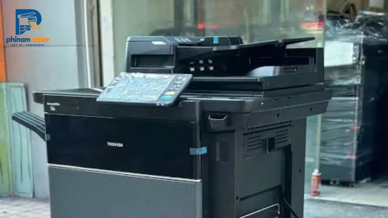 Máy photocopy Toshiba màu đen đặt ngoài trời, có bảng điều khiển cảm ứng và khay giấy.